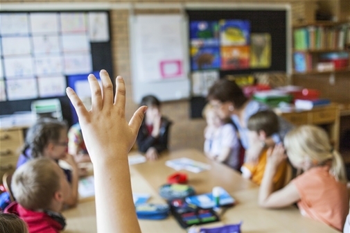 Barnhand räcks upp i förgrunden i klassrum med elever och lärare i bakgrunden